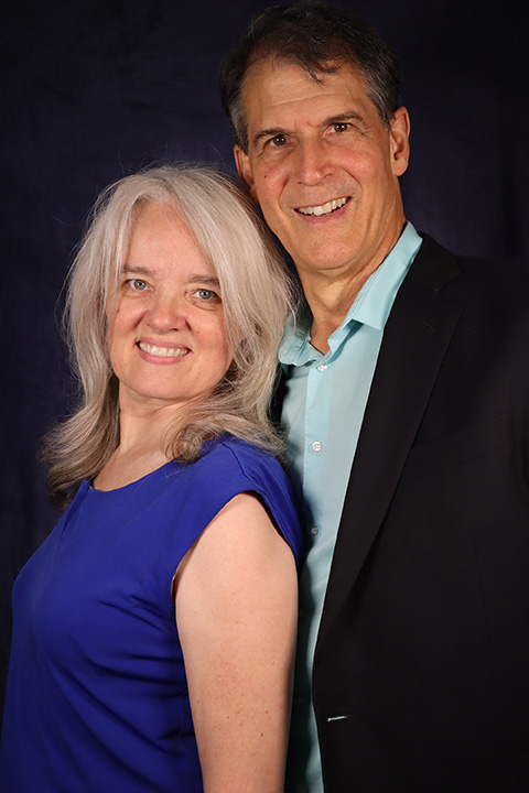 Couple posing for a portrait: woman with silver hair in a blue dress and man in a teal shirt and black blazer, against a dark backdrop.