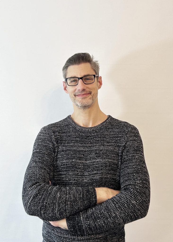 Man with short gray hair and glasses, crossing his arms and smiling slightly in a light, neutral background wearing a dark gray knit sweater