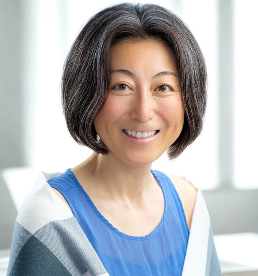 Smiling woman with short dark hair wearing a blue blouse, indoors with soft light in the background.